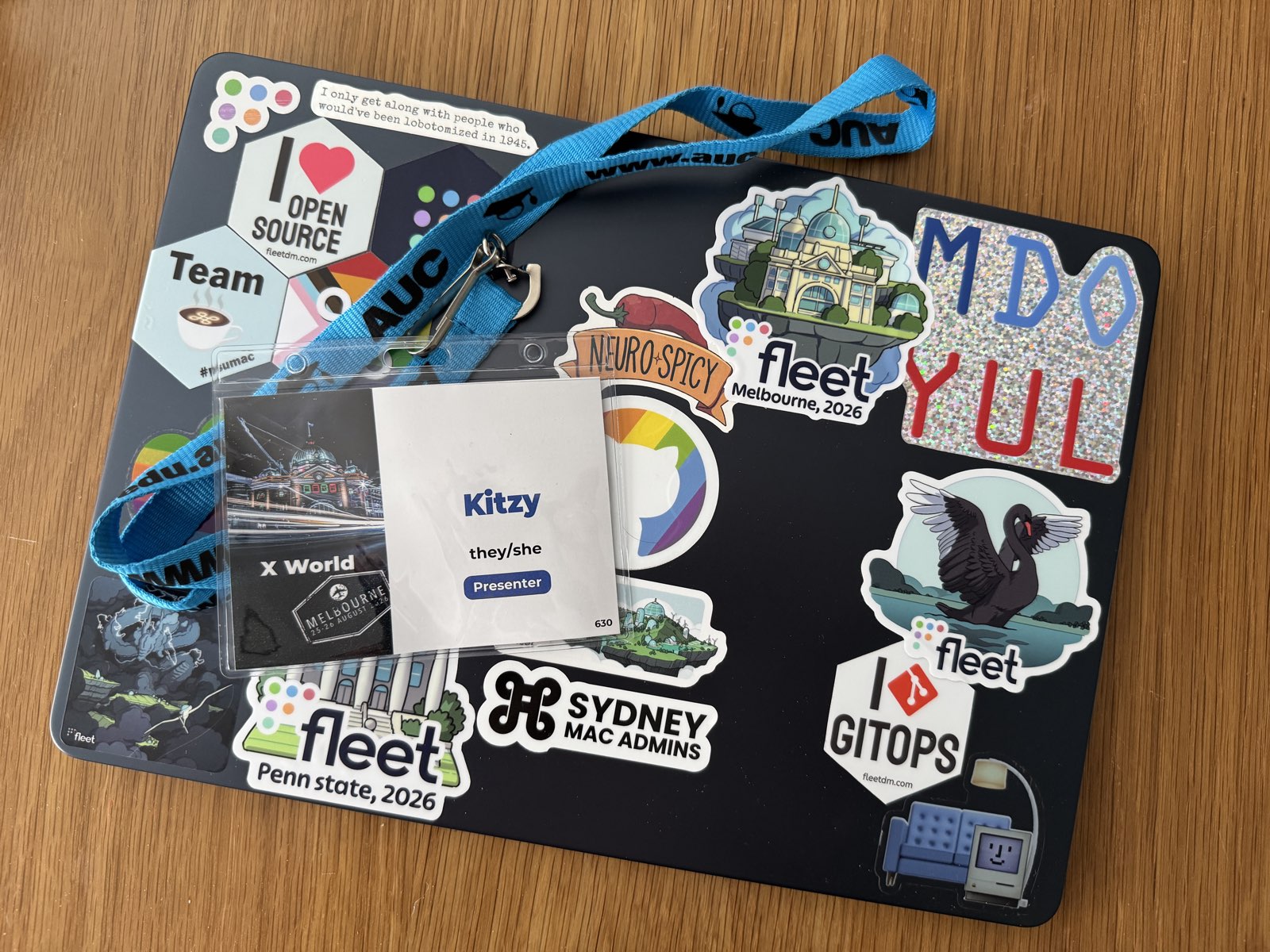 Laptop covered in years of Mac admin community stickers, with an X World 2026 presenter badge reading "Kitzy, they/she" on top